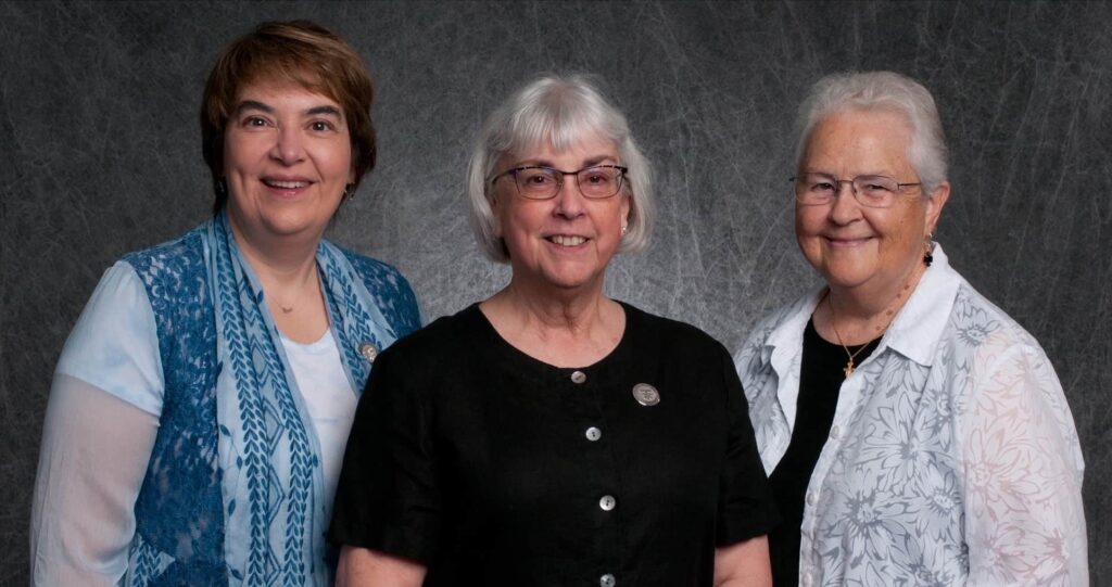 Three women standing in front of a gray background. 